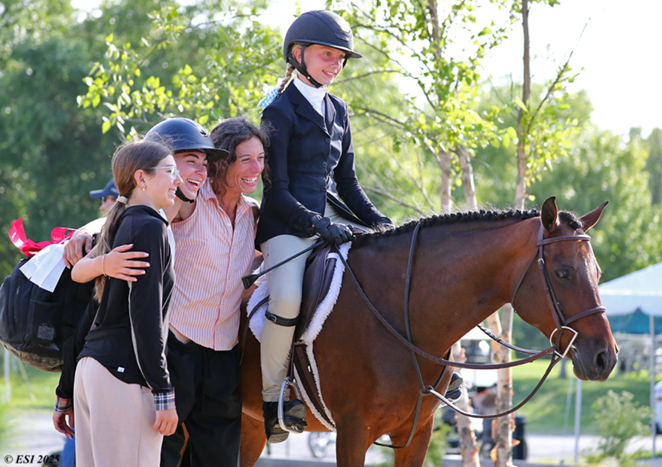 A smiling helmeted rider on a bay horse with three people gathered beside them at a horse show, with the original ESI 2025 credit visible
