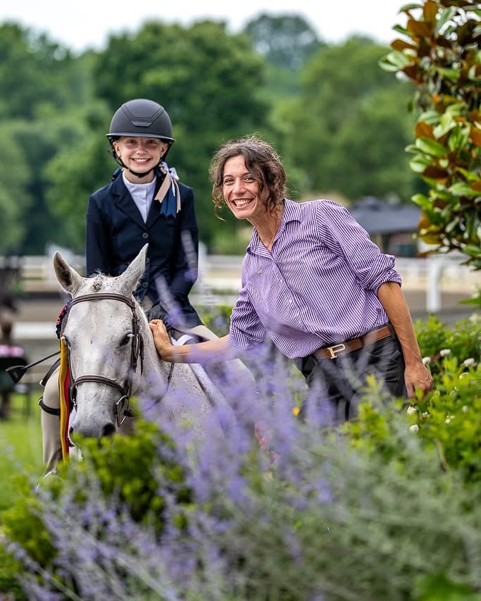 A smiling young helmeted rider on a grey horse beside a smiling woman in a striped shirt, framed by soft purple flowers and green foliage