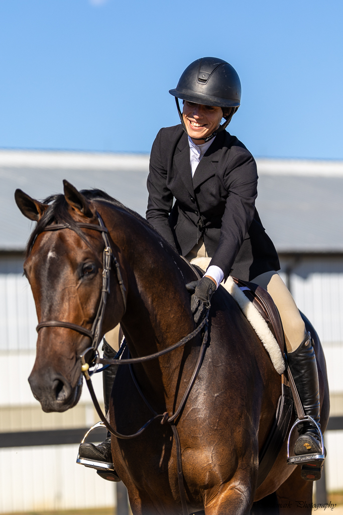 Leah Posner in a black show jacket, white shirt and black helmet, smiling warmly as she sits atop her bay horse Broadway and rests a gloved hand on his neck, with a softly blurred barn behind them