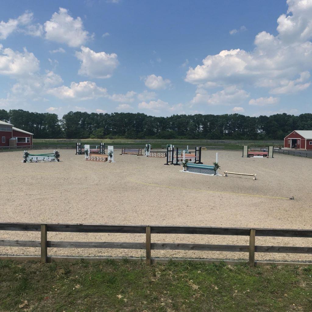 Outdoor hunter-jumper arena with colorful jumps, sandy footing and red barns under a blue sky at Posnerosa