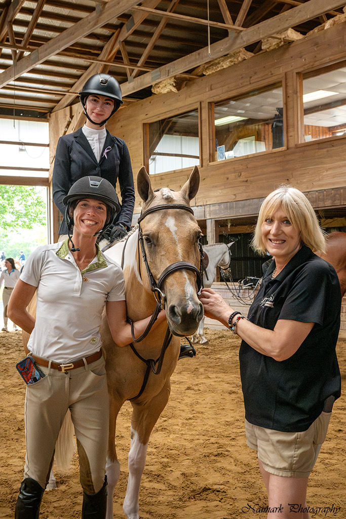 A helmeted rider on a golden horse with two smiling people standing beside its head inside an indoor arena, with the original Naimark Photography credit visible