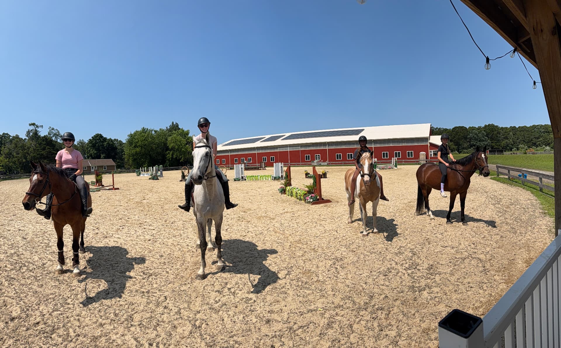 Four helmeted riders on bay, grey and pinto horses in the sunny Posnerosa outdoor arena with the red indoor barn behind them