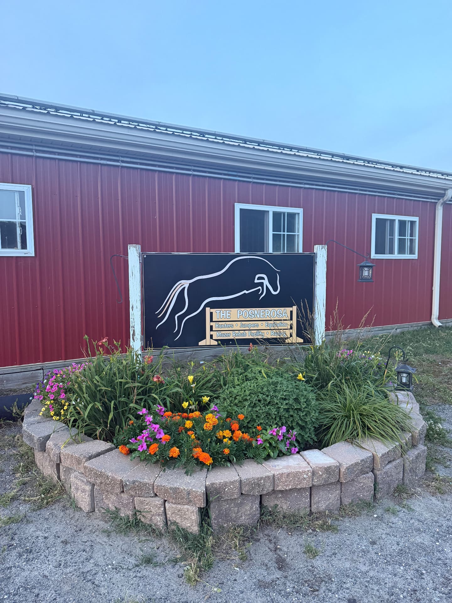 The Posnerosa horse sign beside a red barn and a stone-bordered garden filled with colorful flowers