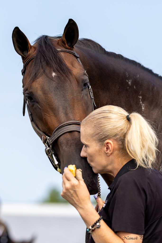 Colleen Posner in profile, gently kissing the muzzle of her dark bay horse Broadway against a soft blue sky, with the original Naimark Photography credit visible