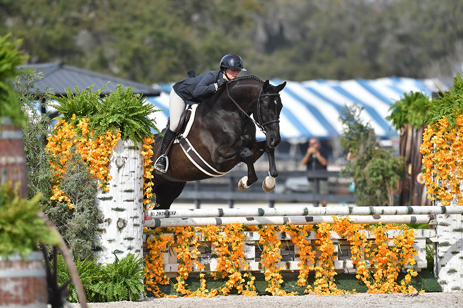 A helmeted rider on a dark horse clearing a white hunter fence decorated with orange flowers and green ferns, with the original ESI 2026 photography credit visible