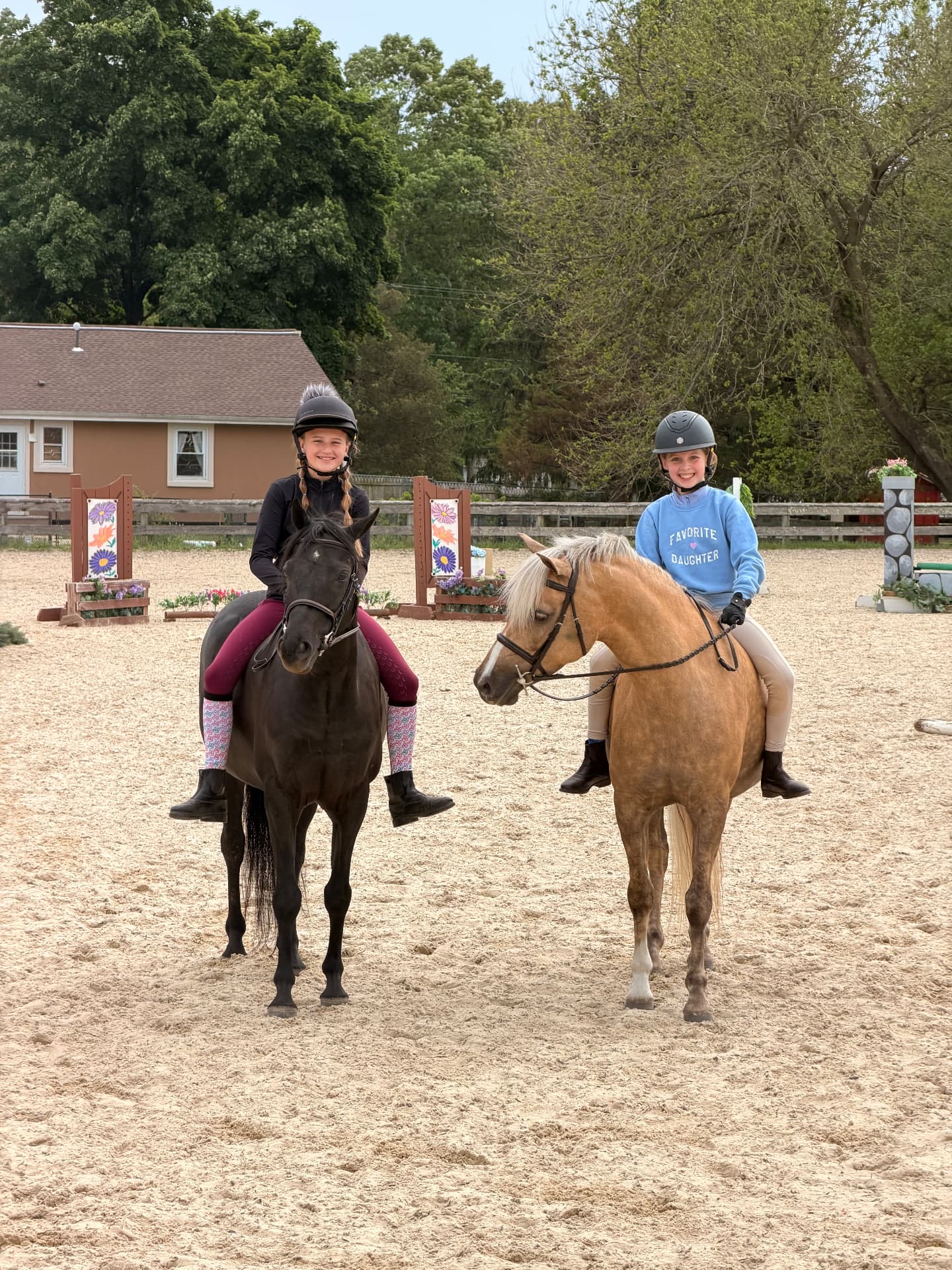 Two smiling helmeted young riders sitting side by side on a dark pony and a golden pony in the Posnerosa outdoor arena