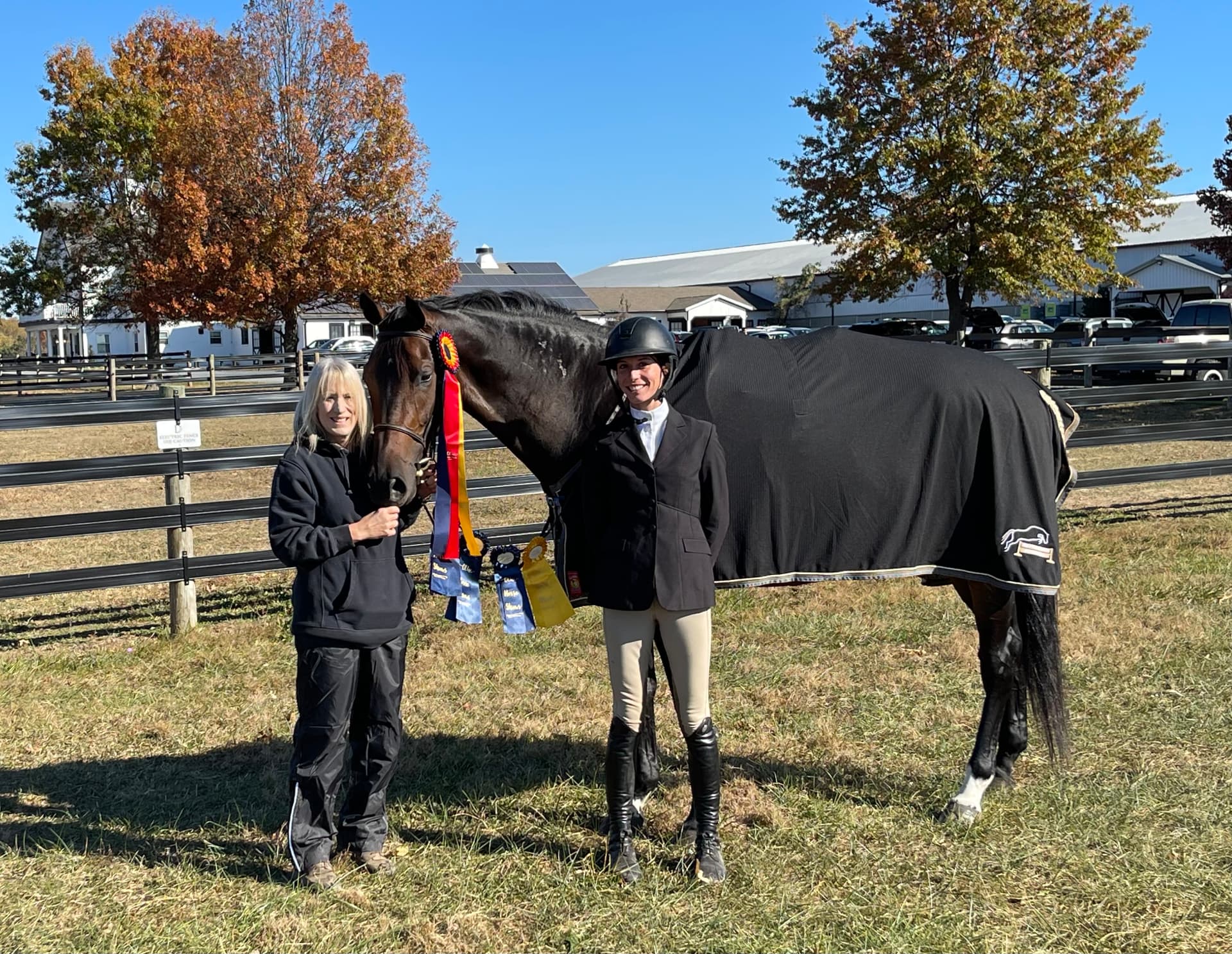 A dark bay horse wearing a black cooler draped with award rosettes, standing beside a smiling rider in show attire and a person holding its bridle in an autumn paddock with fall foliage