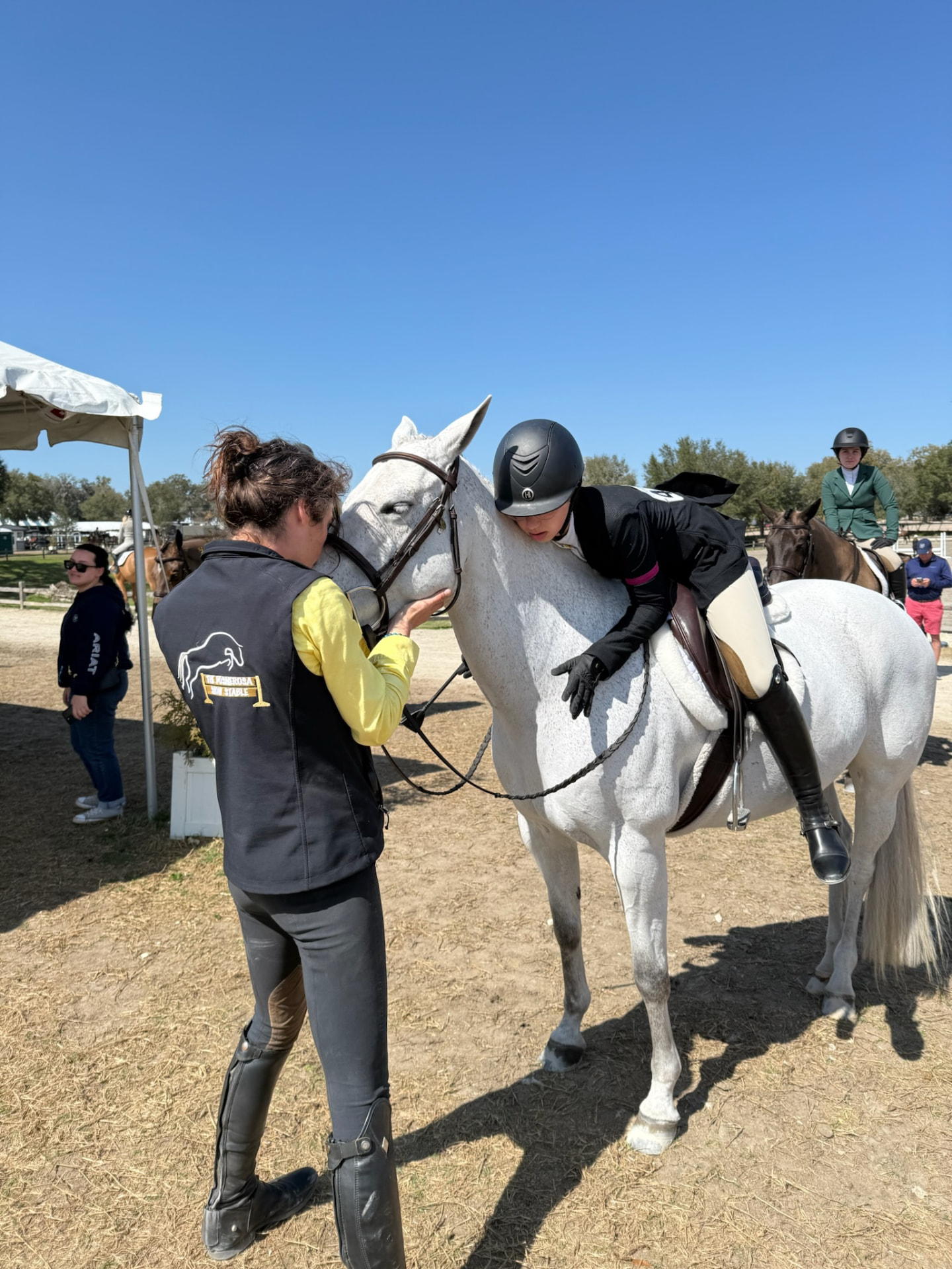 A helmeted rider leaning close to a grey horse while a person wearing a Posnerosa vest stands beside its head at a horse show