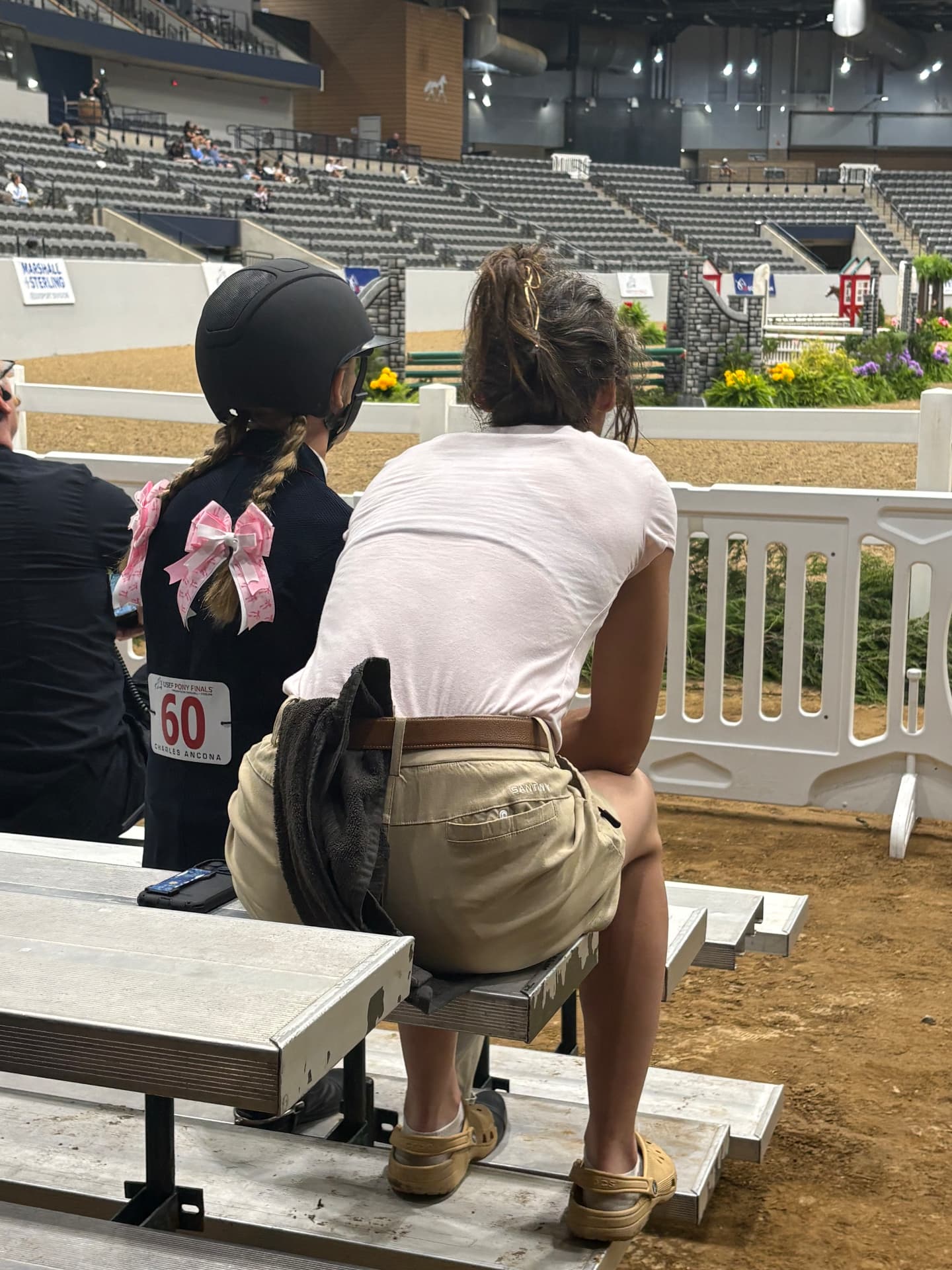A young helmeted rider with pink hair bows and an adult watching an indoor horse-show arena together from the ringside benches